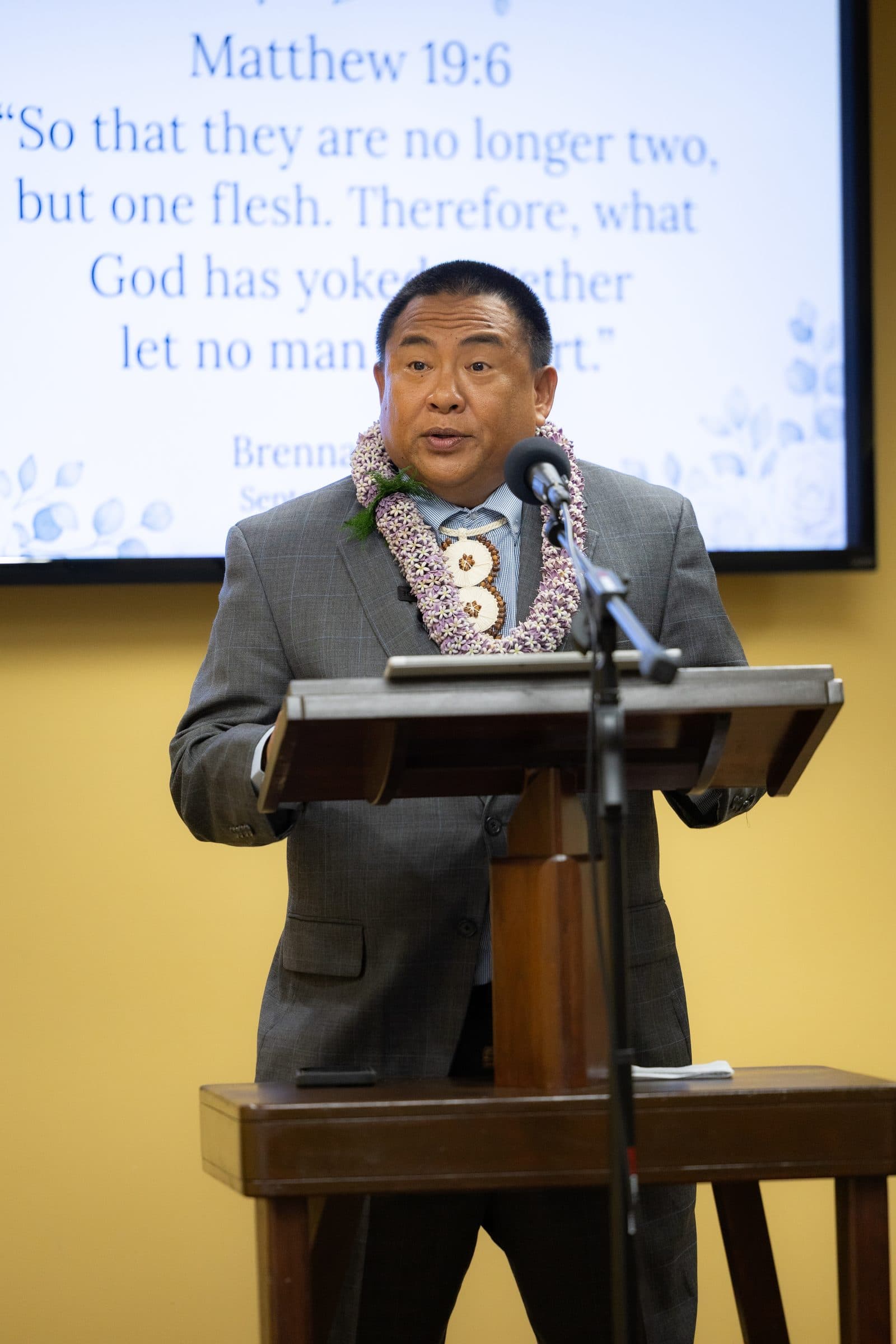 The officiant, in lei, speaks at a lectern beneath a projected Bible verse.
