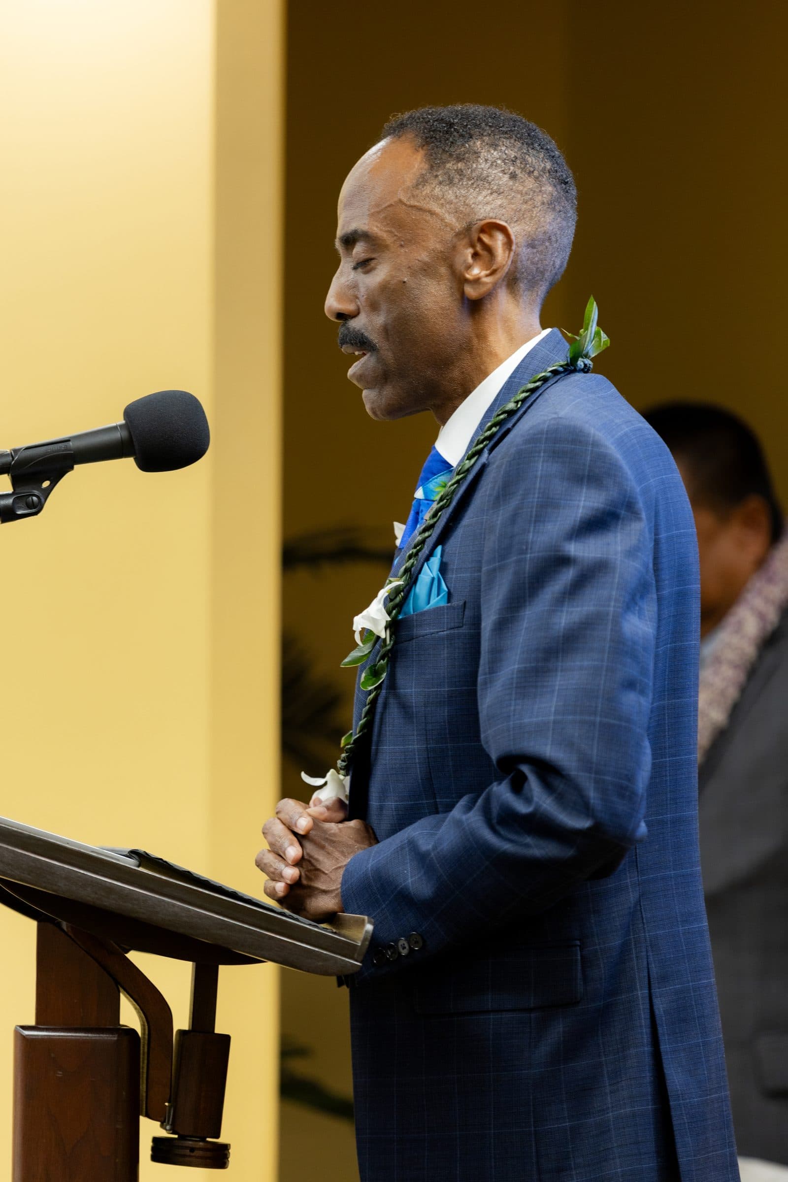 A man in a blue jacket and lei speaks at the microphone.