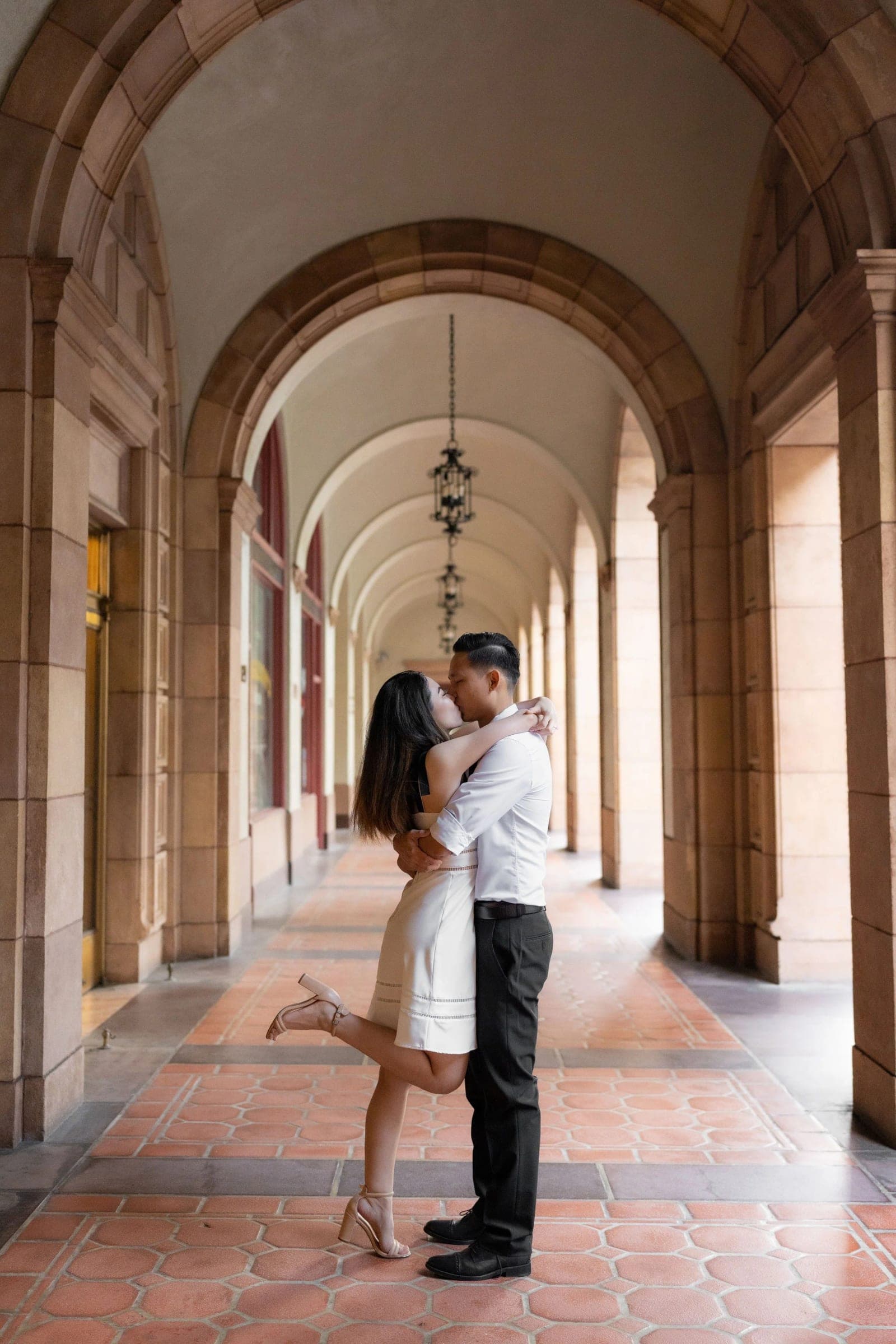 A couple kiss in a long arched stone walkway, warm light on the tiles.