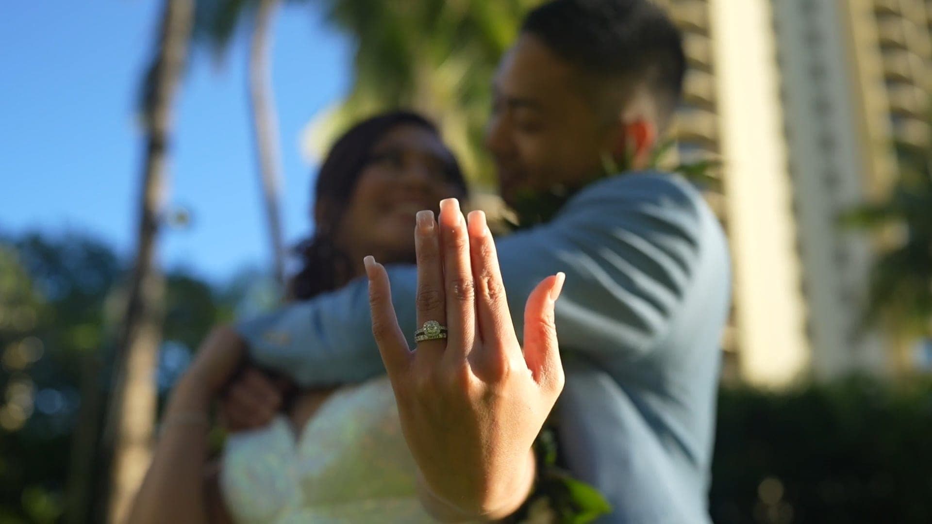 Ann Mirae shows her ring to the camera as Nathan hugs her, palms and towers behind.