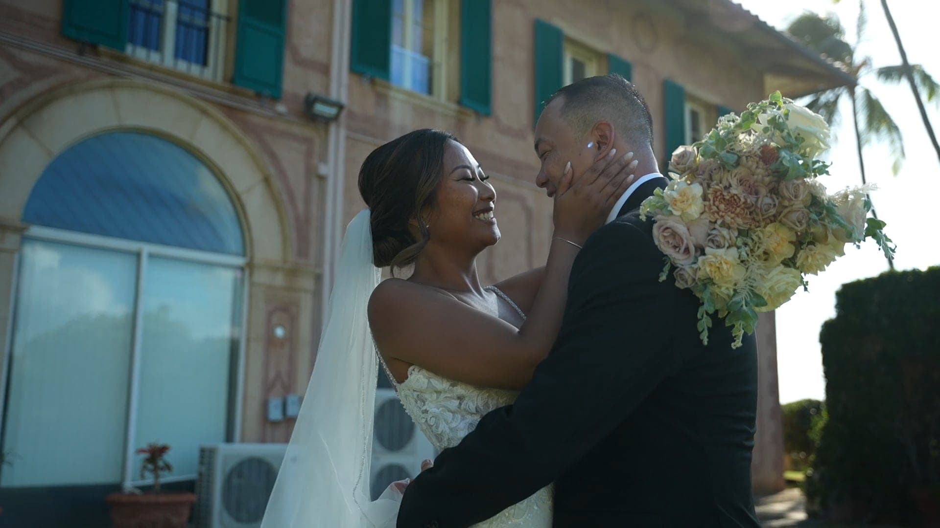 Lesley holds Johnny's face and laughs, her bouquet over his shoulder, outside a pink building.