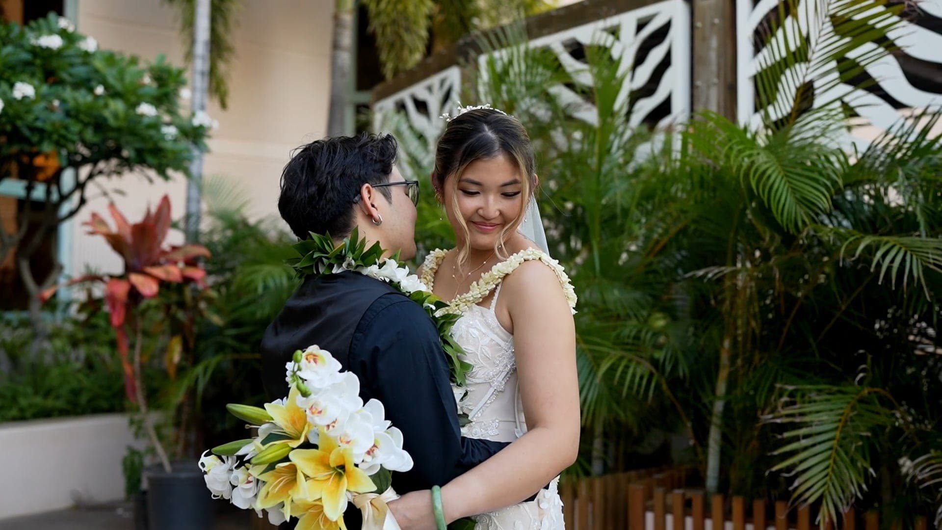 Matthew and Sarah, in lei and holding white flowers, smile together beside a garden of palms.