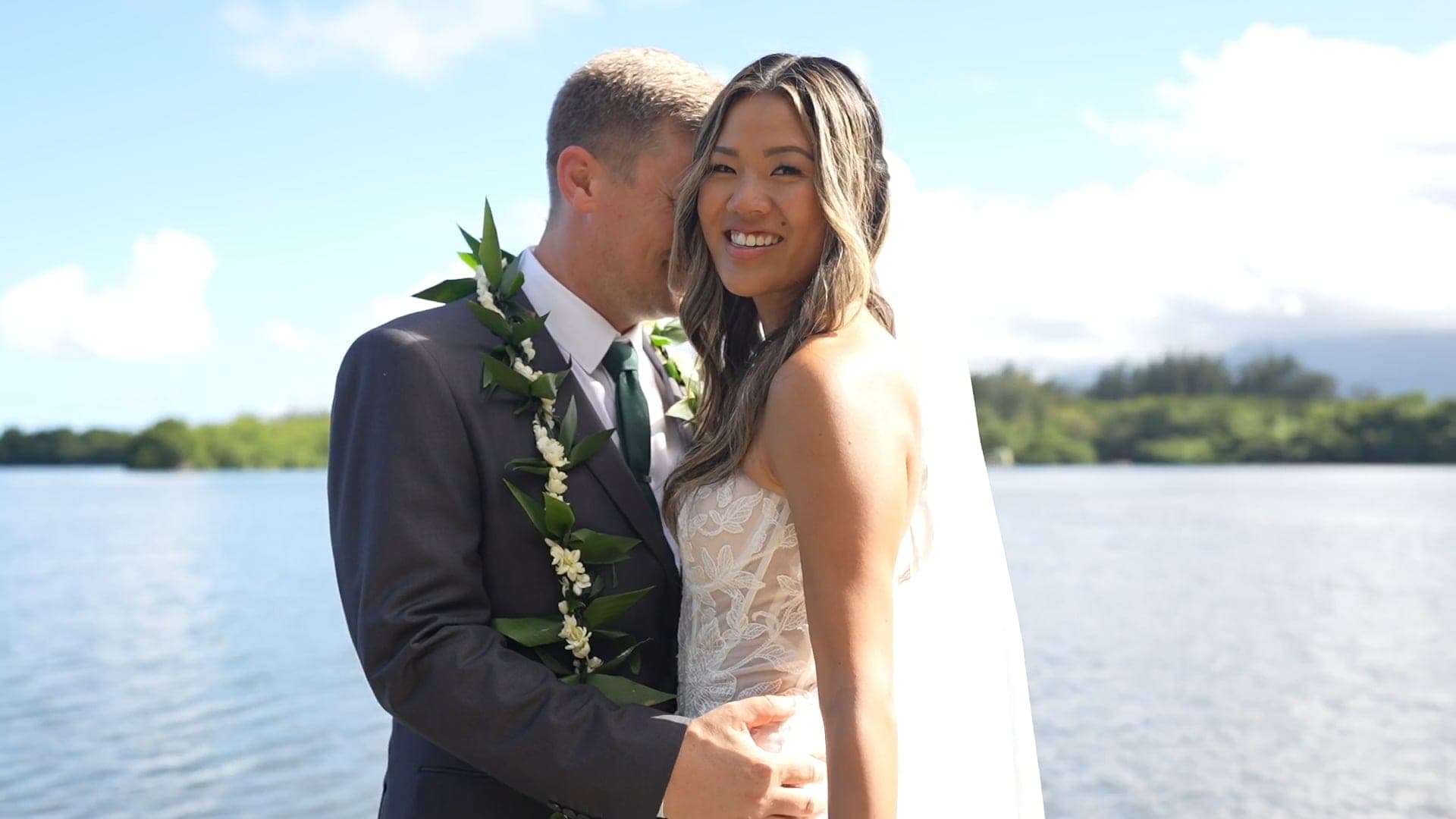 Arlan, in a green leaf lei, leans close as Stacey smiles at the camera beside calm water.