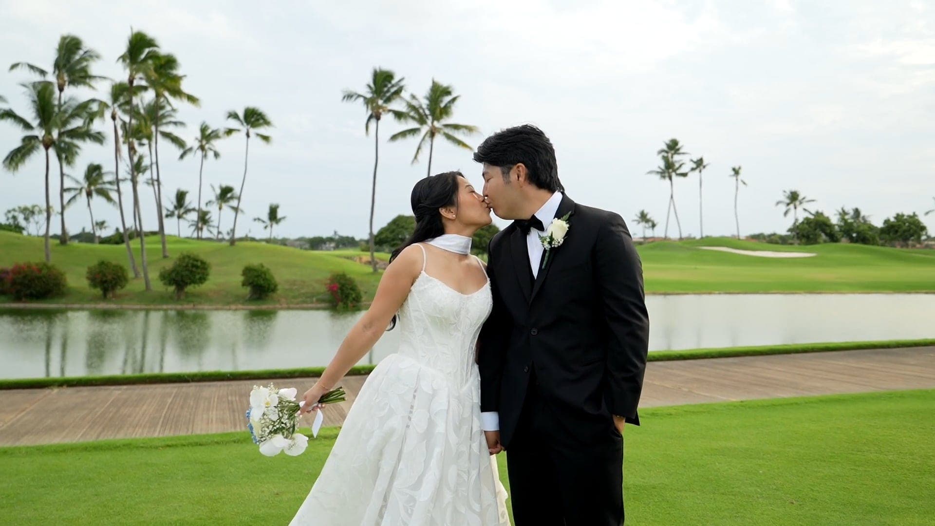 Mia and Brennan kiss on a golf course lawn lined with palm trees.