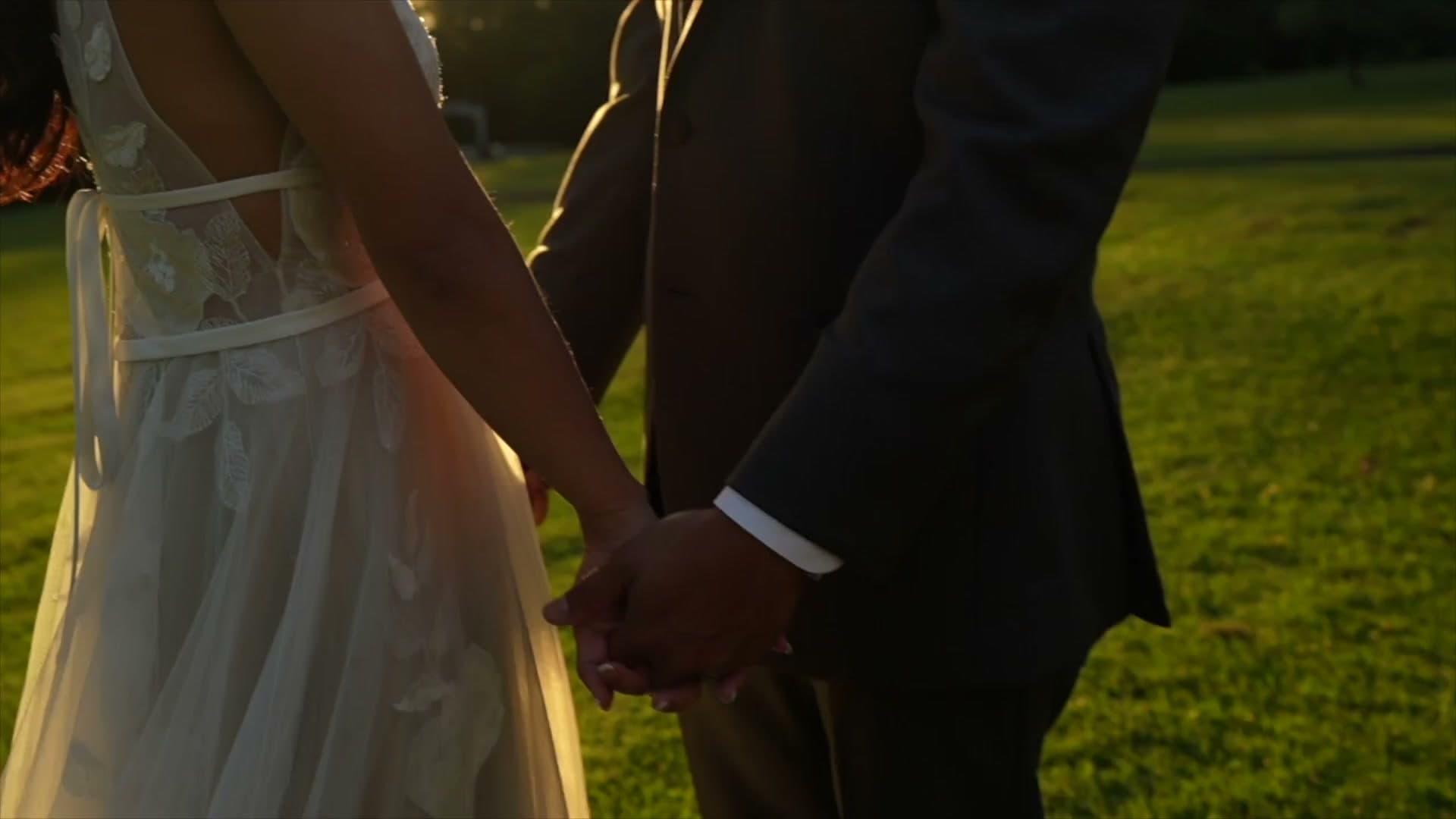 A bride and groom hold hands as they walk across a lawn in golden evening light.