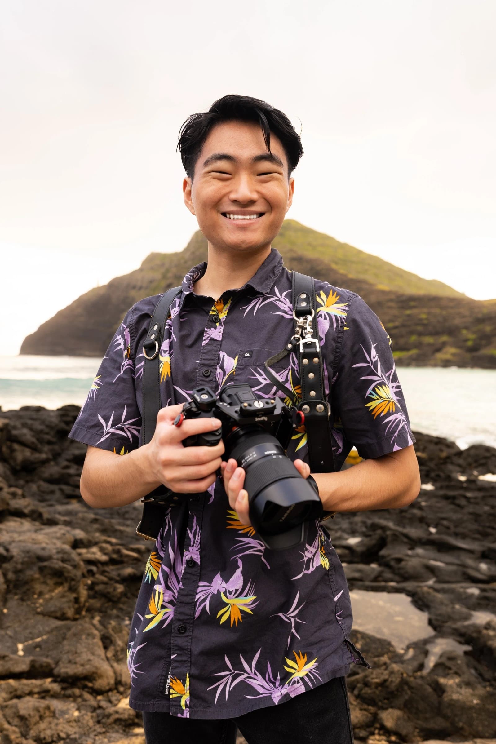 Justin smiling with his camera on a rocky stretch of coast.